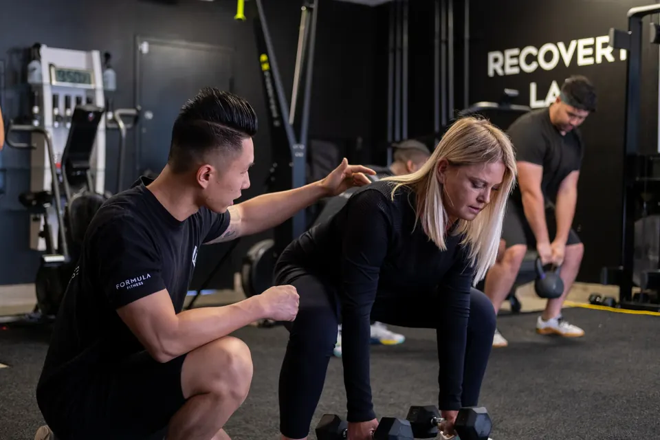 A Formula Fitness coach crouches beside a client who lowers two dumbbells in a hinge position, with another client behind